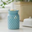 Hobnail Blue wax melt warmer sits on a table beside a white, ceramic butter dish with a lid and a green, potted plant in the background